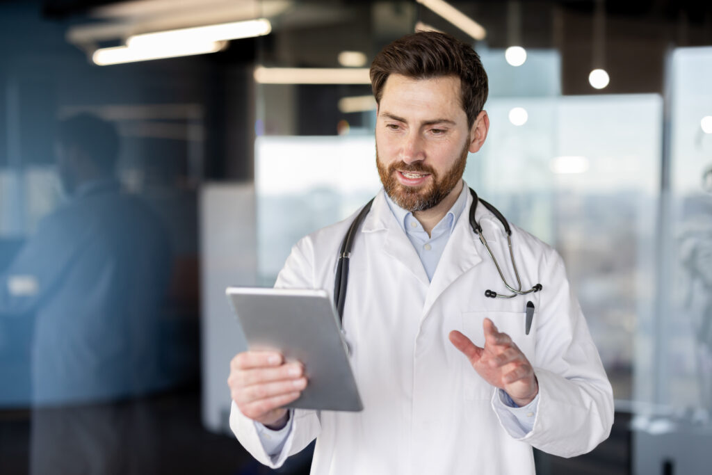 Doctor joven con bata blanca y estetoscopio utilizando una tablet en una oficina moderna, simbolizando el uso de tecnología en telemedicina.
