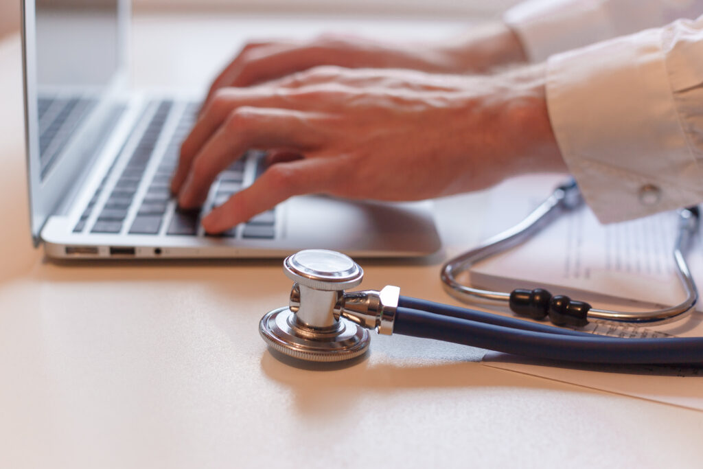 Médico escribiendo en su laptop con fonendoscopio sobre la mesa, representando la emisión de licencias médicas online.
